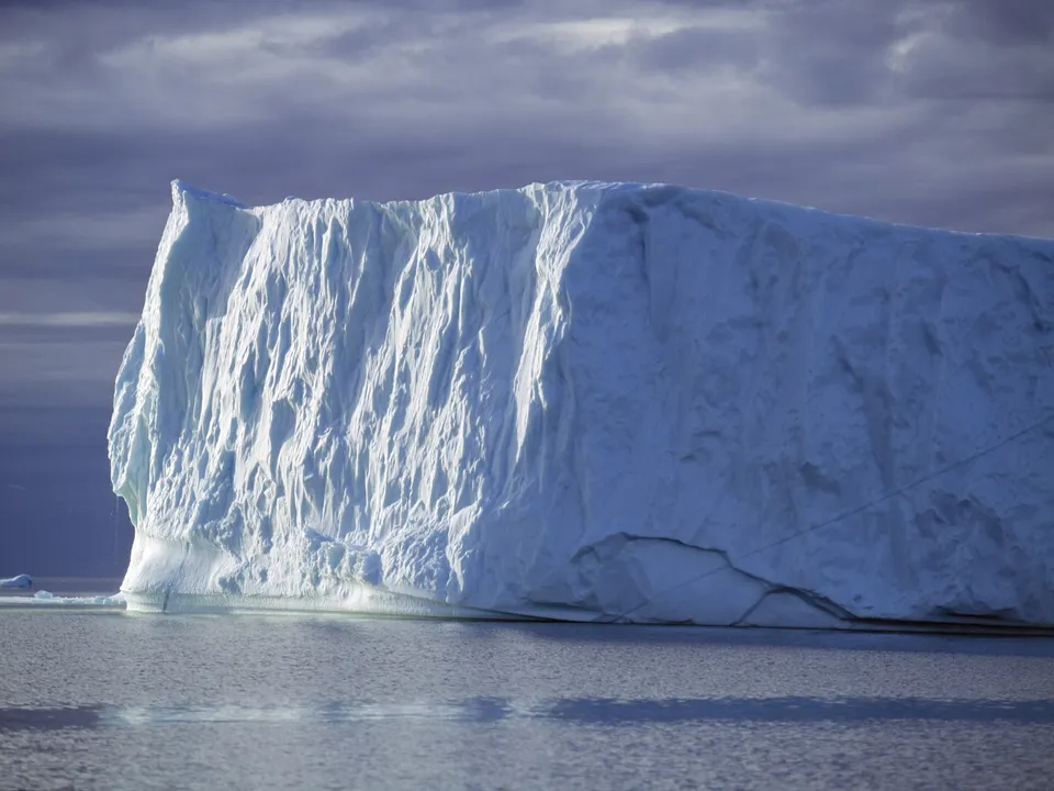 A huge iceberg with a textured surface reflects in calm water beneath a dramatic, cloudy sky.