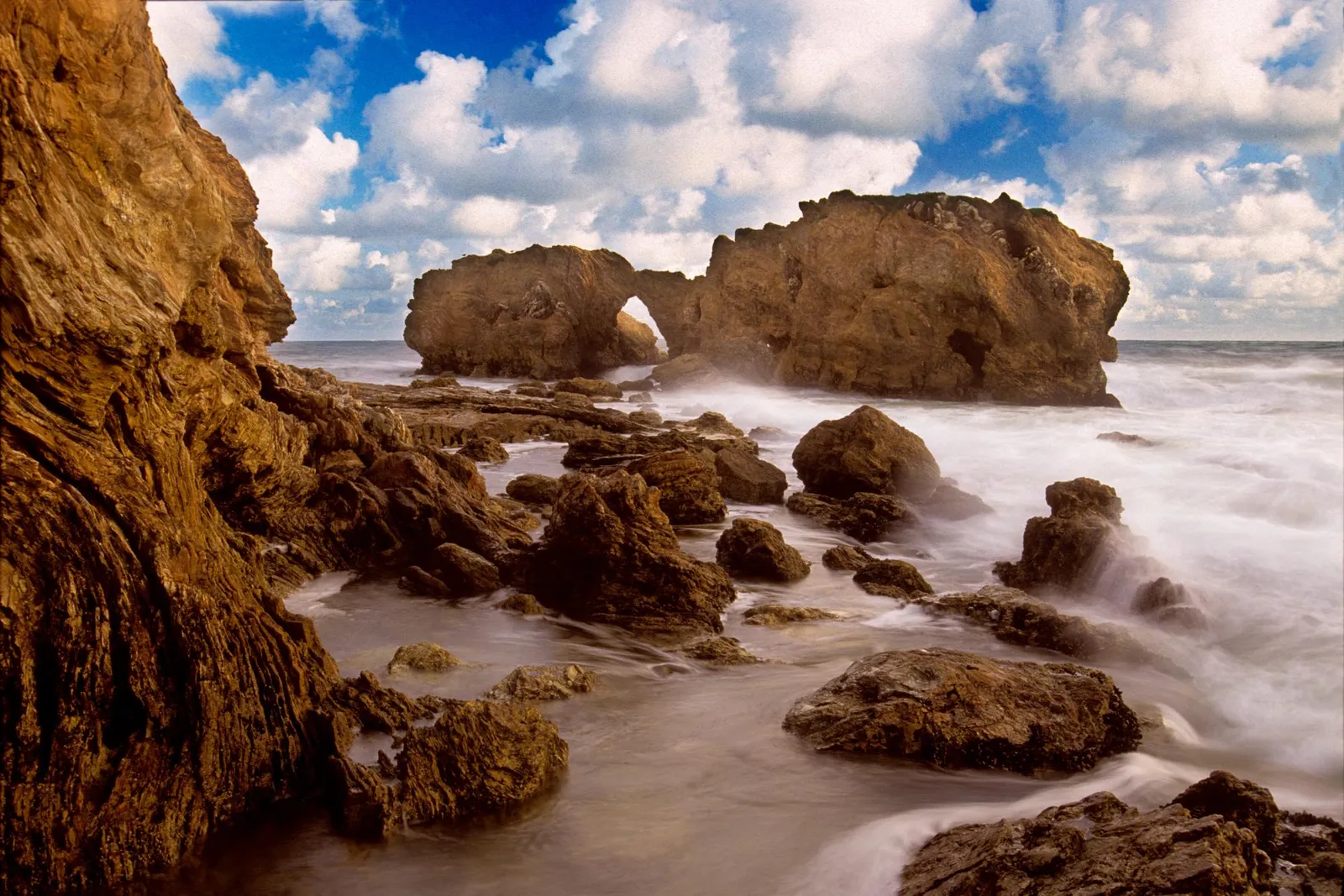 A rocky coastline with waves crashing against large rock formations and an arch.