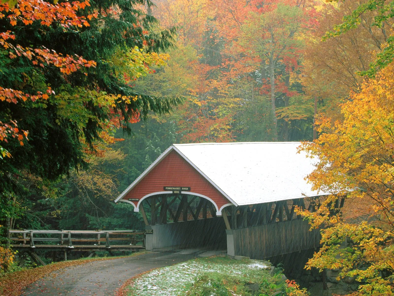 A red covered bridge with a white roof sits amidst trees displaying vibrant autumn colors and a dusting of snow.
