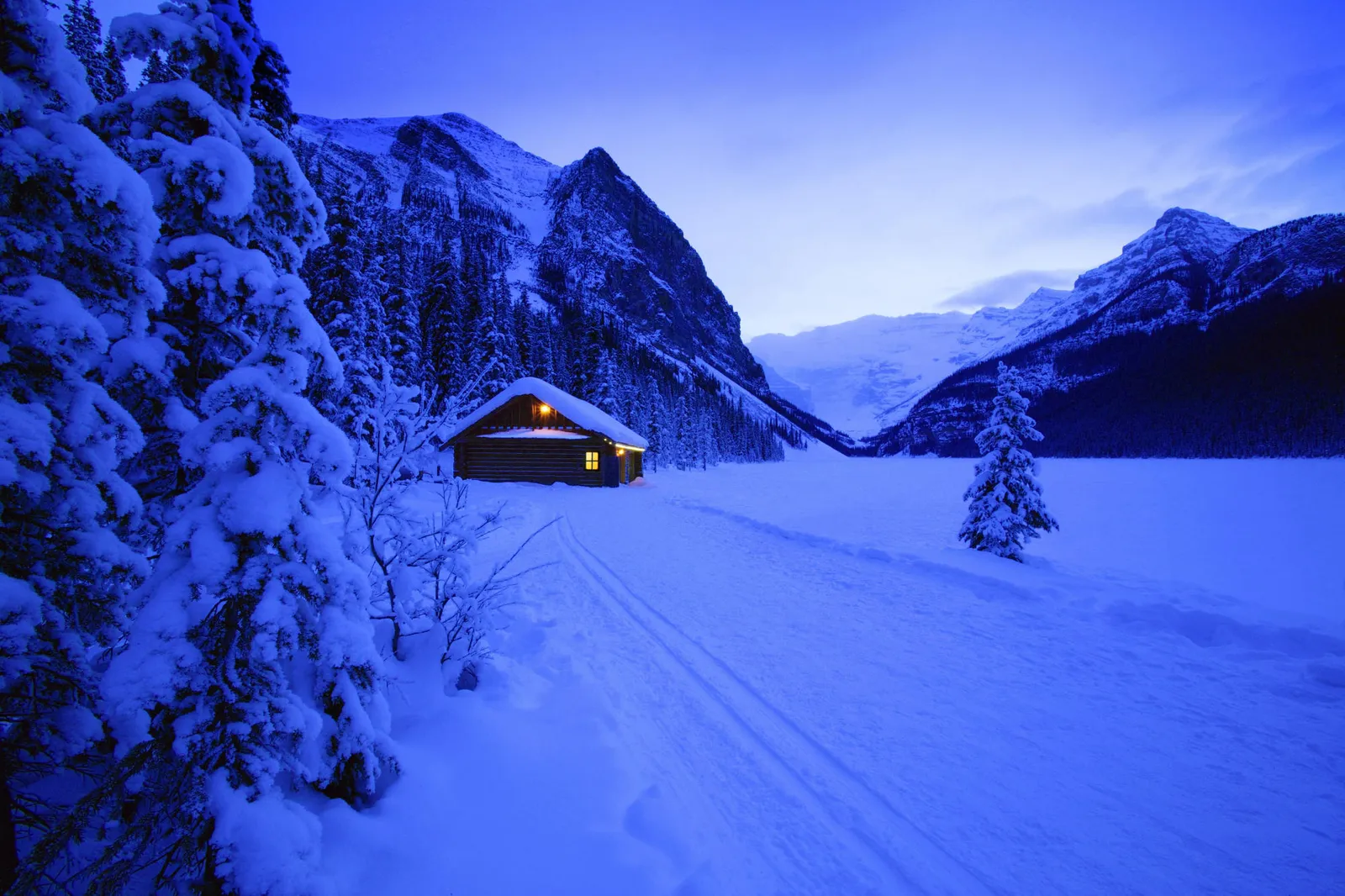 A wooden cabin glows with warm light in a snowy landscape at dusk.