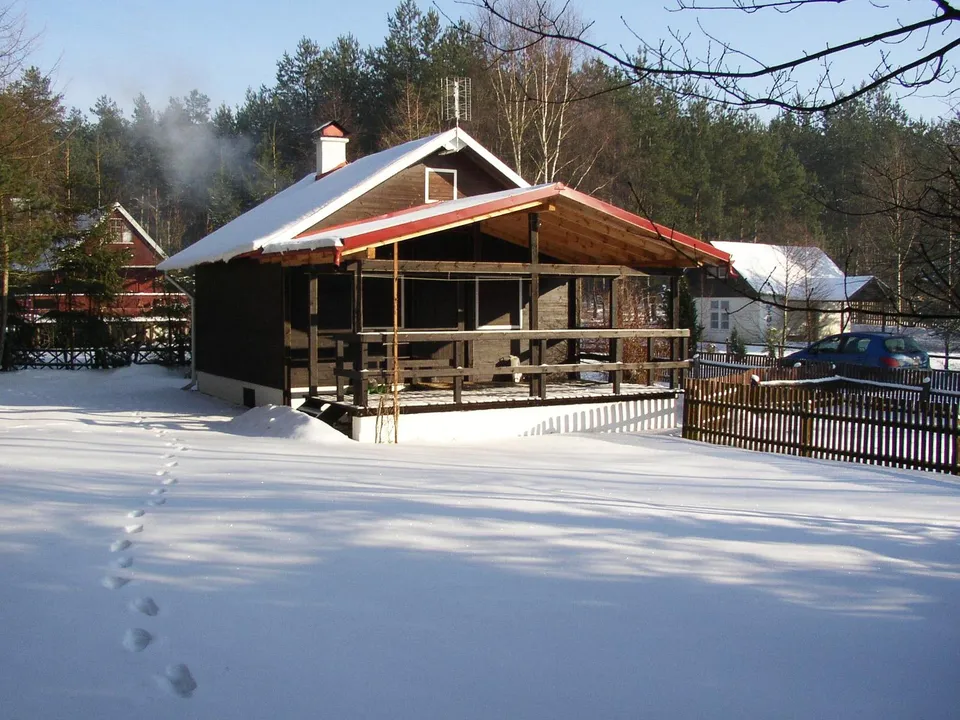 A cozy cabin in a snowy forest has smoke rising from its chimney on a sunny day.