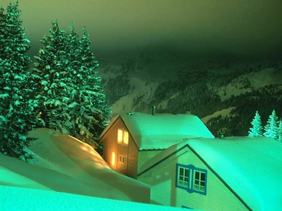 Two cabins are covered in snow at night, with warm light coming from the windows.