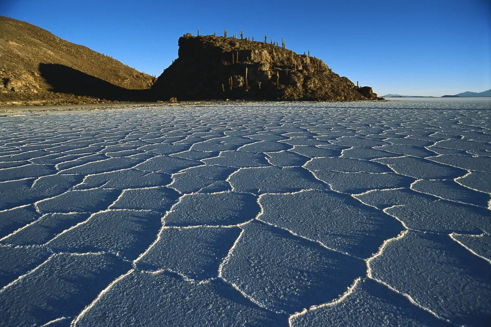 A vast expanse of cracked salt flats stretches towards a rocky island with cacti under a clear blue sky.