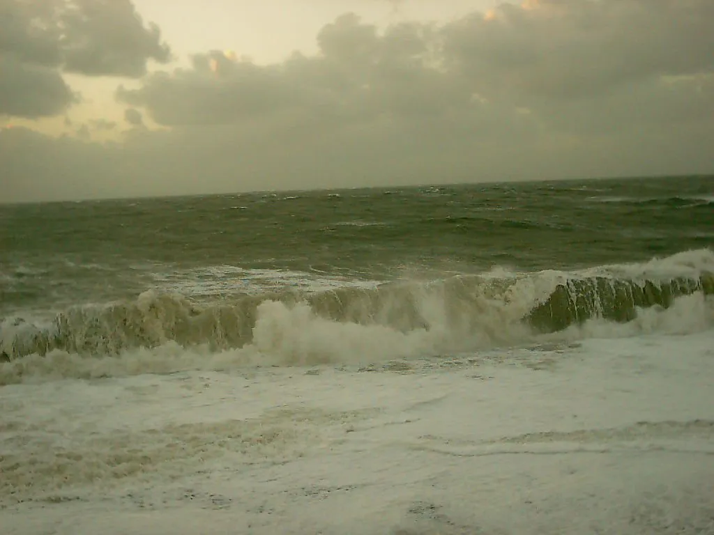 A large wave crashes onto the shore with white foam and spray against a cloudy sky.