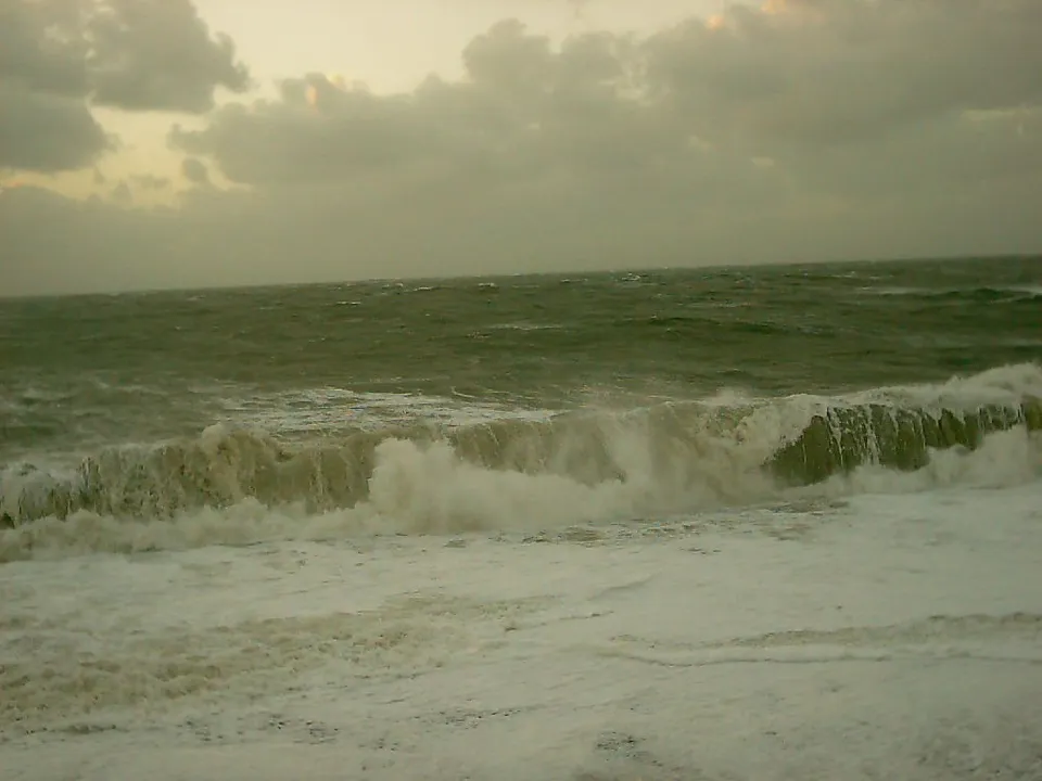A large wave crashes onto the shore with white foam and spray against a cloudy sky.