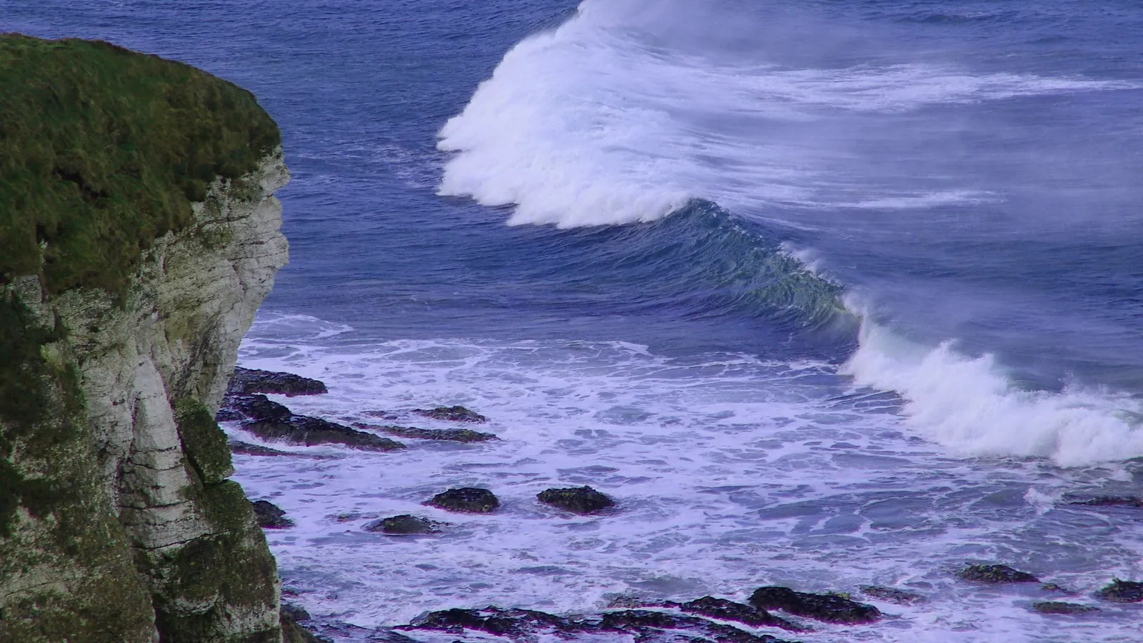 A large wave crashes against a rugged, grassy cliff face overlooking the ocean.
