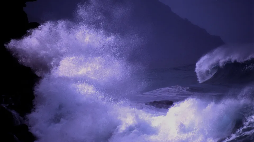 Large waves crash violently against a dark, rocky coastline under a deep blue sky.