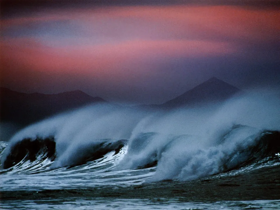 Powerful ocean waves crash with spray against a backdrop of silhouetted mountains and a colorful sunset sky.
