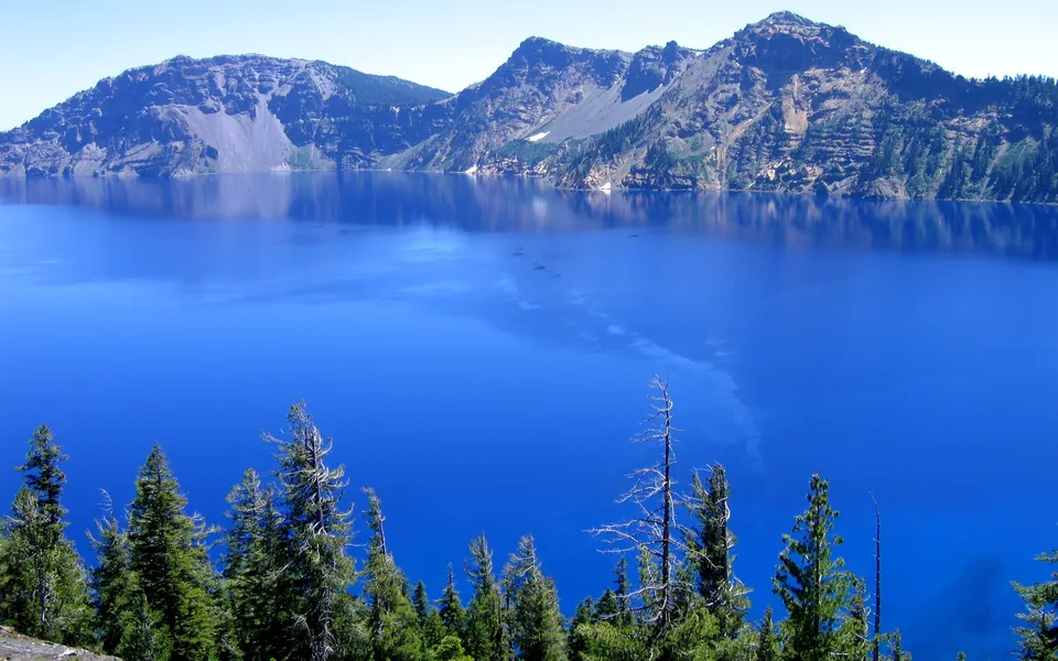 A wide, deep blue crater lake is bordered by rugged, tree-covered mountains under a clear sky.