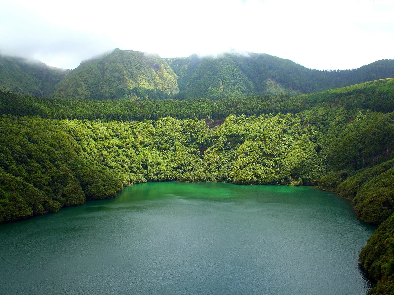 A wide view of a deep green crater lake surrounded by steep, densely forested green mountains under a cloudy sky.