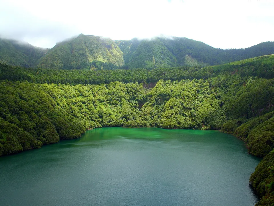 A wide view of a deep green crater lake surrounded by steep, densely forested green mountains under a cloudy sky.
