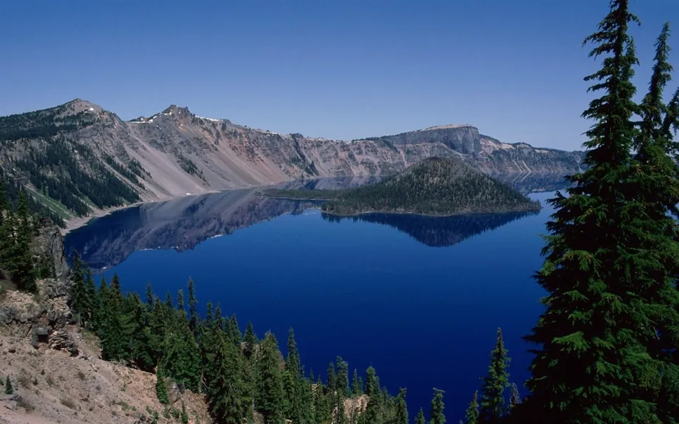 A wide view of a deep blue lake surrounded by mountains and evergreen trees, with calm water reflecting the scenery.