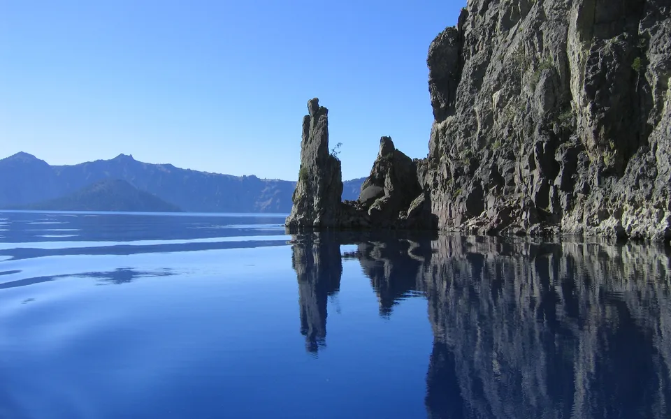 Tall rock spires rise from a deep blue lake, with distant mountains and clear blue sky.