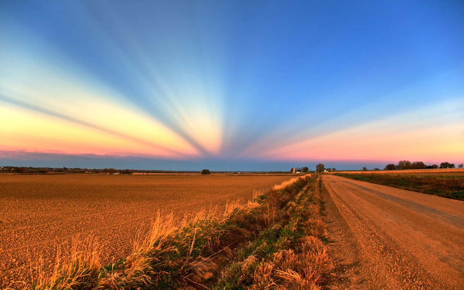Sun rays fan out across a colorful sky above a rural landscape with a dirt road.