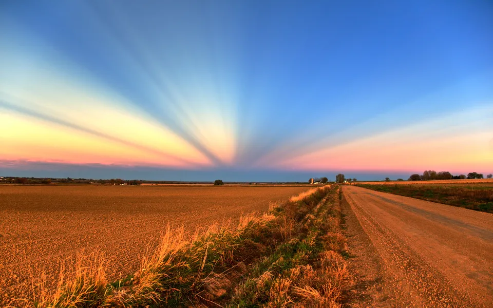Sun rays fan out across a colorful sky above a rural landscape with a dirt road.