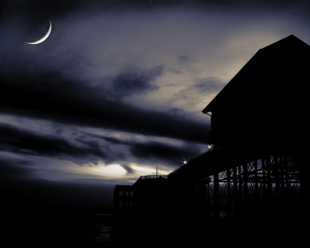 A sliver of a crescent moon hangs in a cloudy night sky above silhouetted buildings and a pier.