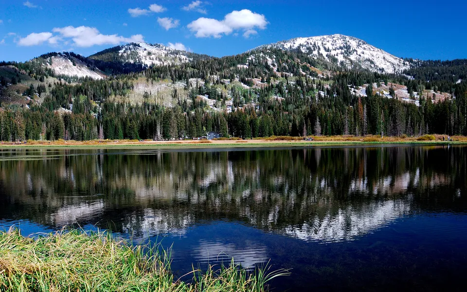 A bright blue sky with clouds reflects in a mountain lake with snow-capped peaks.