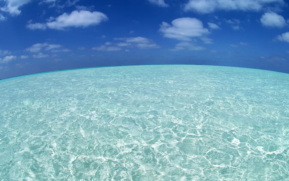 A wide-angle view of shallow, crystal-clear turquoise water under a bright blue sky with scattered white clouds.