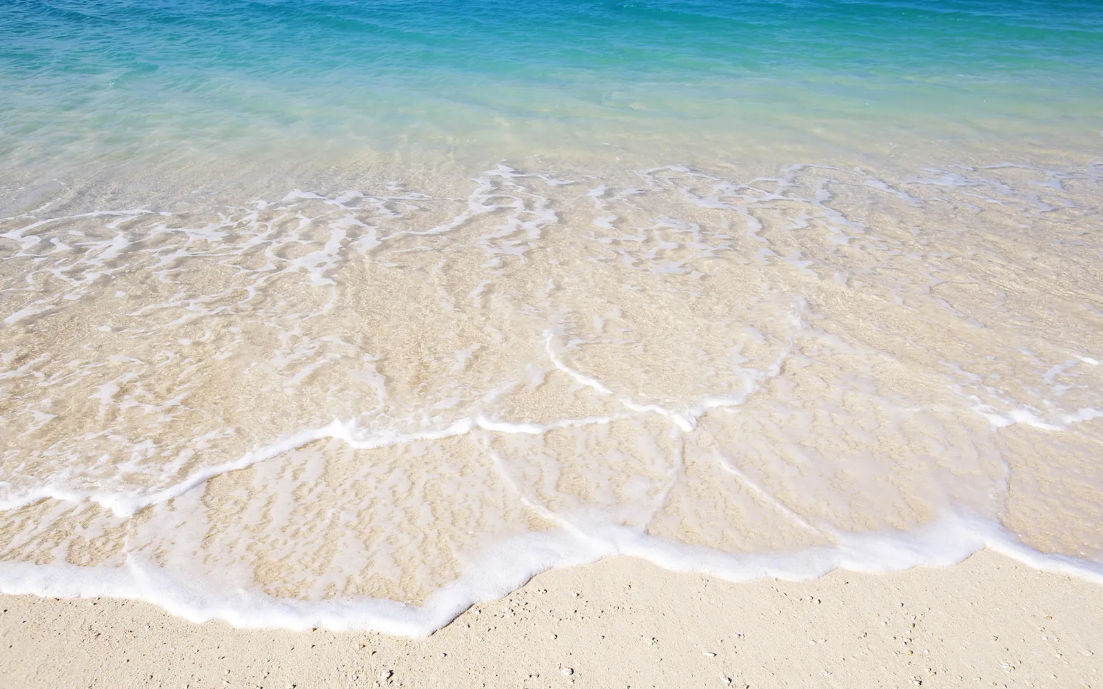 Crystal clear turquoise water gently washes over a light sandy beach, creating delicate patterns of foam.