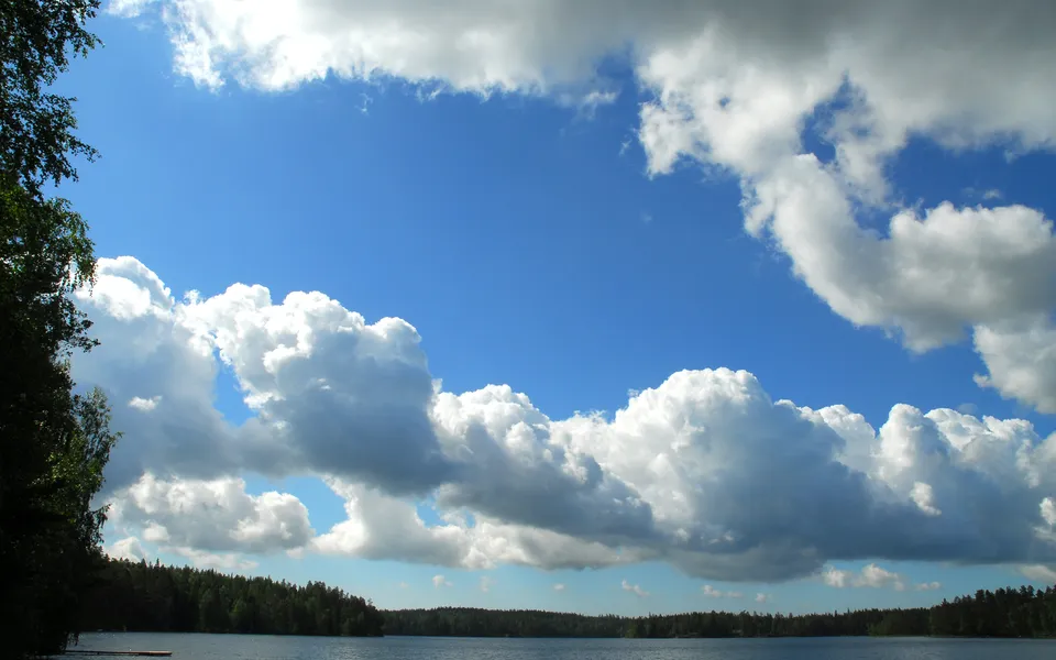 Large, fluffy cumulus clouds fill the bright blue sky above a dark forest and a calm lake.