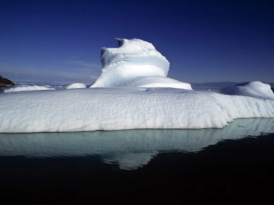 A large iceberg with a curved top floats in dark, calm water, reflecting its form.