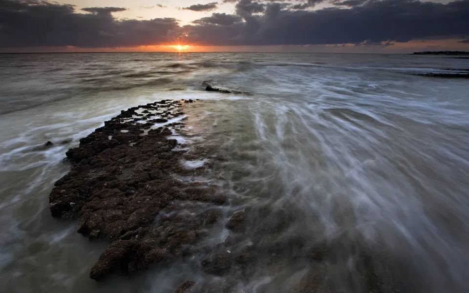 The sun sets over a dark, rocky outcrop on the coastline with waves flowing around it.