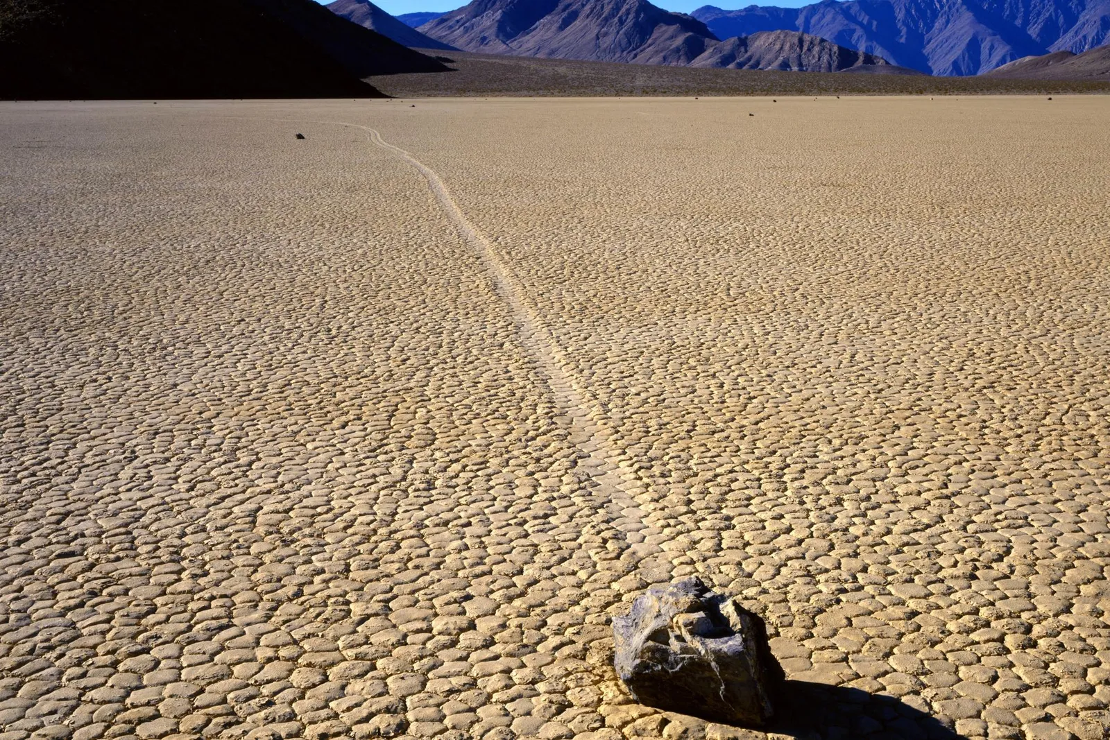 A single rock has left a long, winding track across a cracked, dry lake bed with mountains in the background.