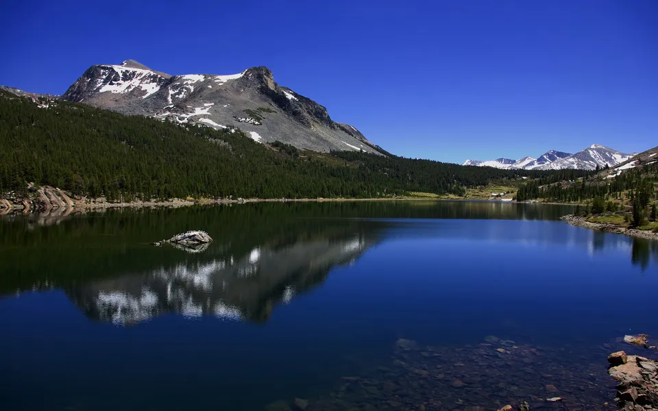 A deep blue lake mirrors snow-capped mountains and evergreen forests under a clear blue sky.