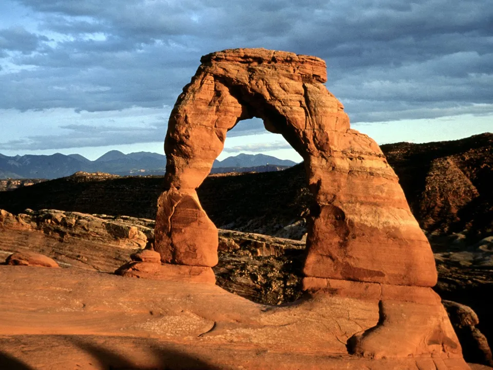 A large sandstone arch glows in the warm light of the setting sun, with distant mountains under a cloudy sky.