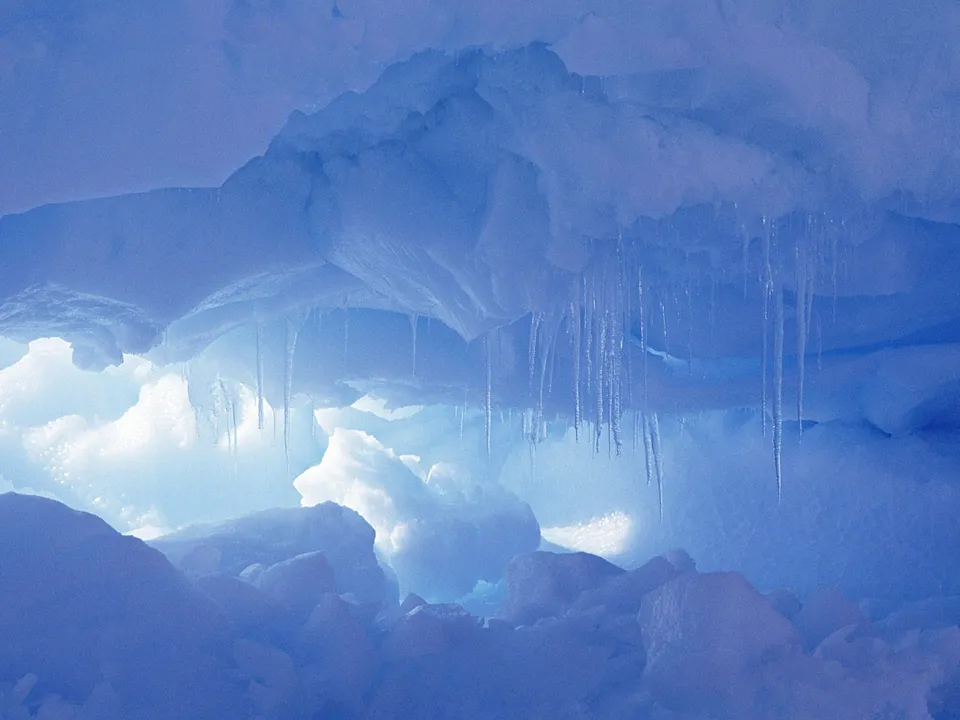 Delicate icicles hang from the ceiling of a bright blue ice cave, illuminated by diffused light.
