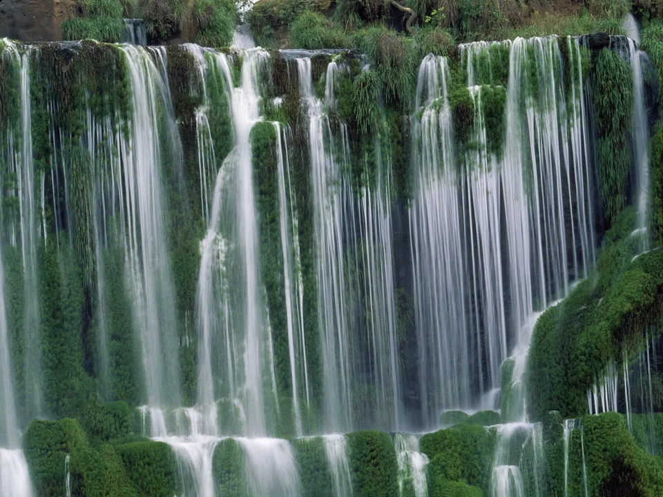 Thin streams of water create a curtain effect over a lush, green cliff face.