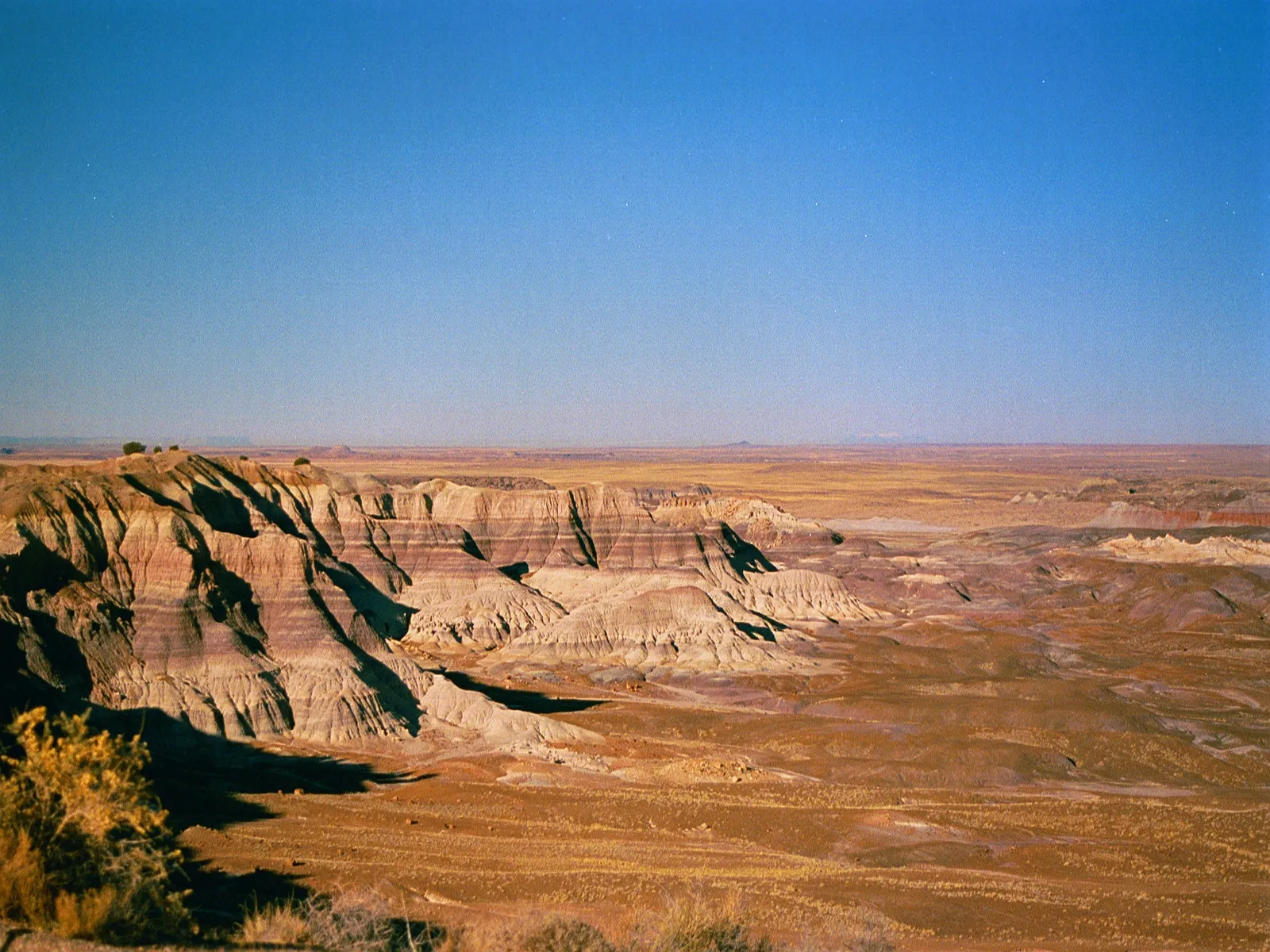 A wide view of colorful desert badlands with layered rock formations under a clear blue sky.