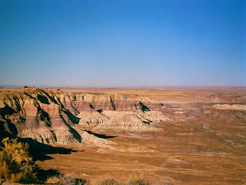 A wide view of colorful desert badlands with layered rock formations under a clear blue sky.