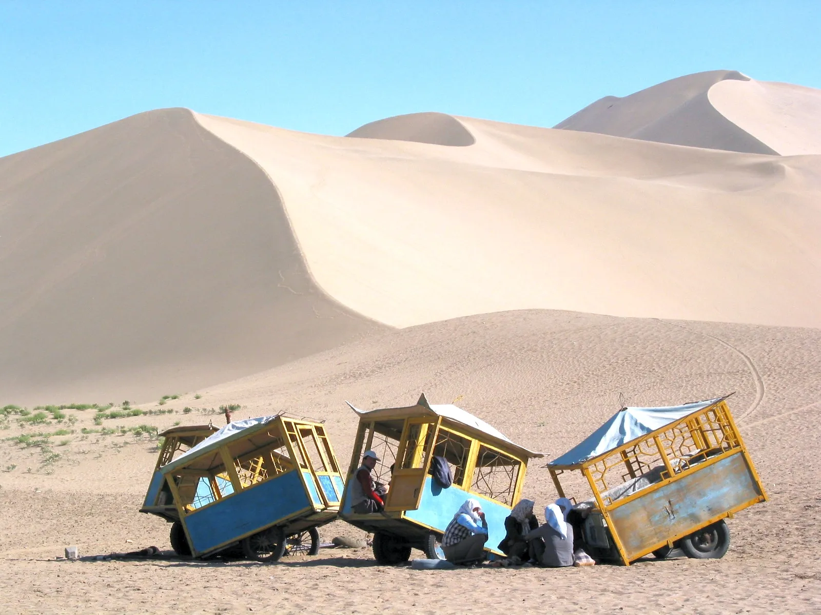 Three colorful tourist carts are parked on a sandy desert landscape with large sand dunes in the background under a clear blue sky.