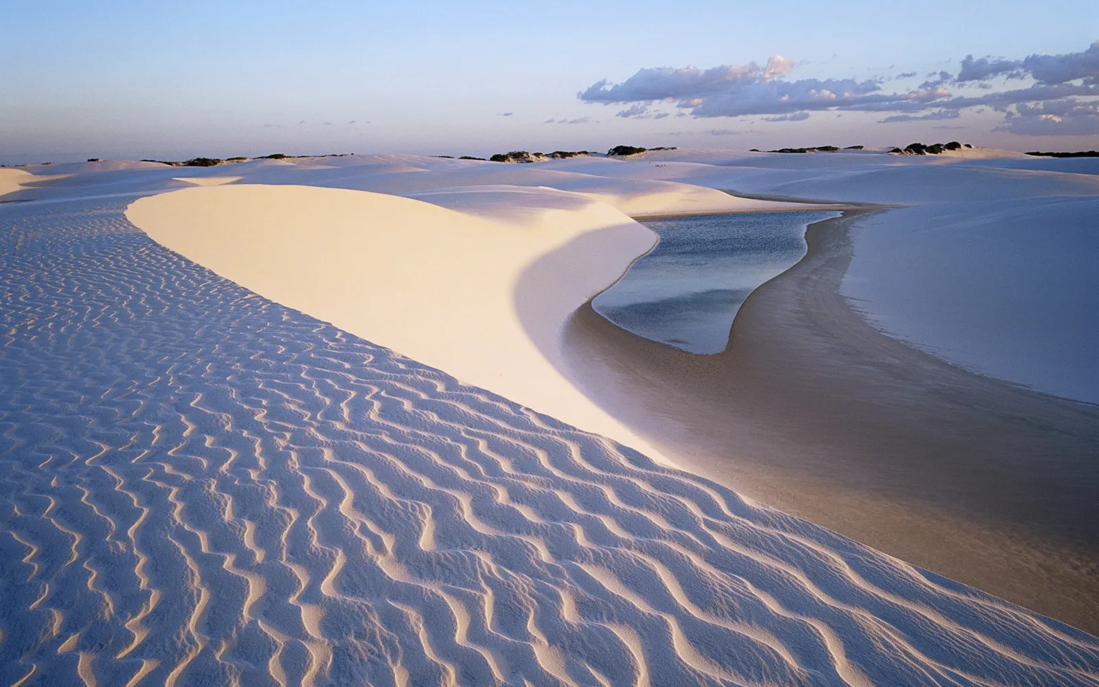 Gentle sand dunes with rippled textures, a small body of water reflecting the sky, and distant vegetation under a clear sky.