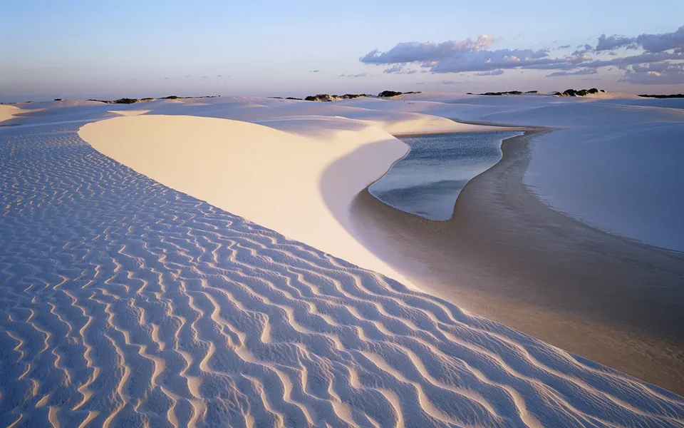 Gentle sand dunes with rippled textures, a small body of water reflecting the sky, and distant vegetation under a clear sky.