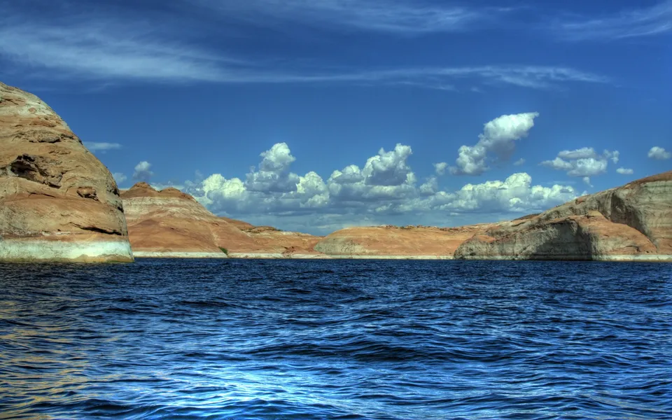 A wide blue lake with rippling water is bordered by dramatic sandstone cliffs under a bright blue sky with scattered clouds.