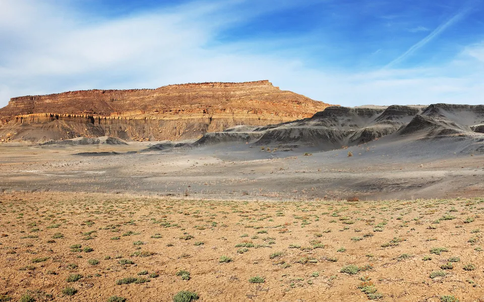 A wide view of a desert with layered rock formations and sparse vegetation under a bright blue sky with wispy clouds.