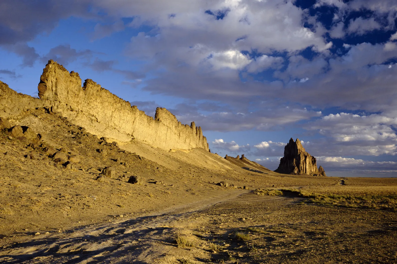 A vast desert landscape with a prominent rock formation and a dirt road leading towards it under a blue sky with clouds.