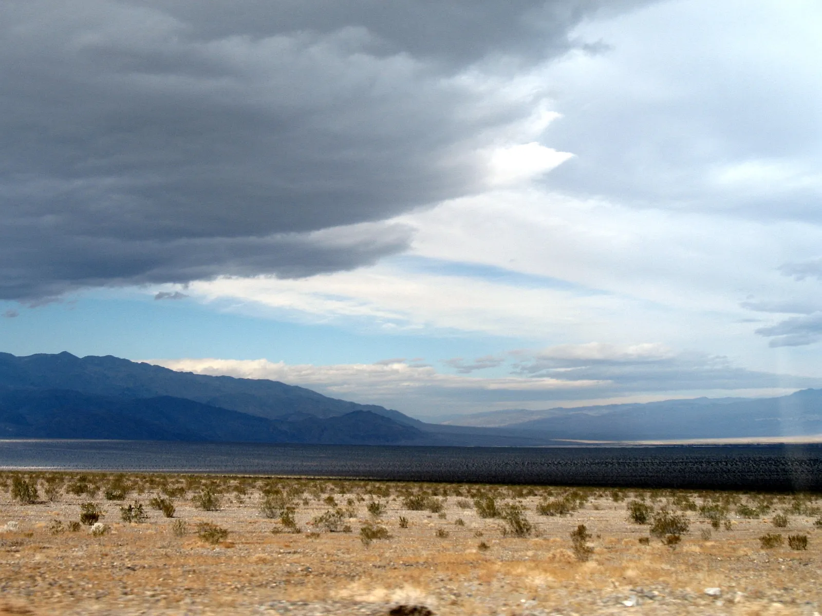 A wide desert landscape with sparse vegetation, dark mountains in the distance, and a dramatic sky filled with dark clouds.