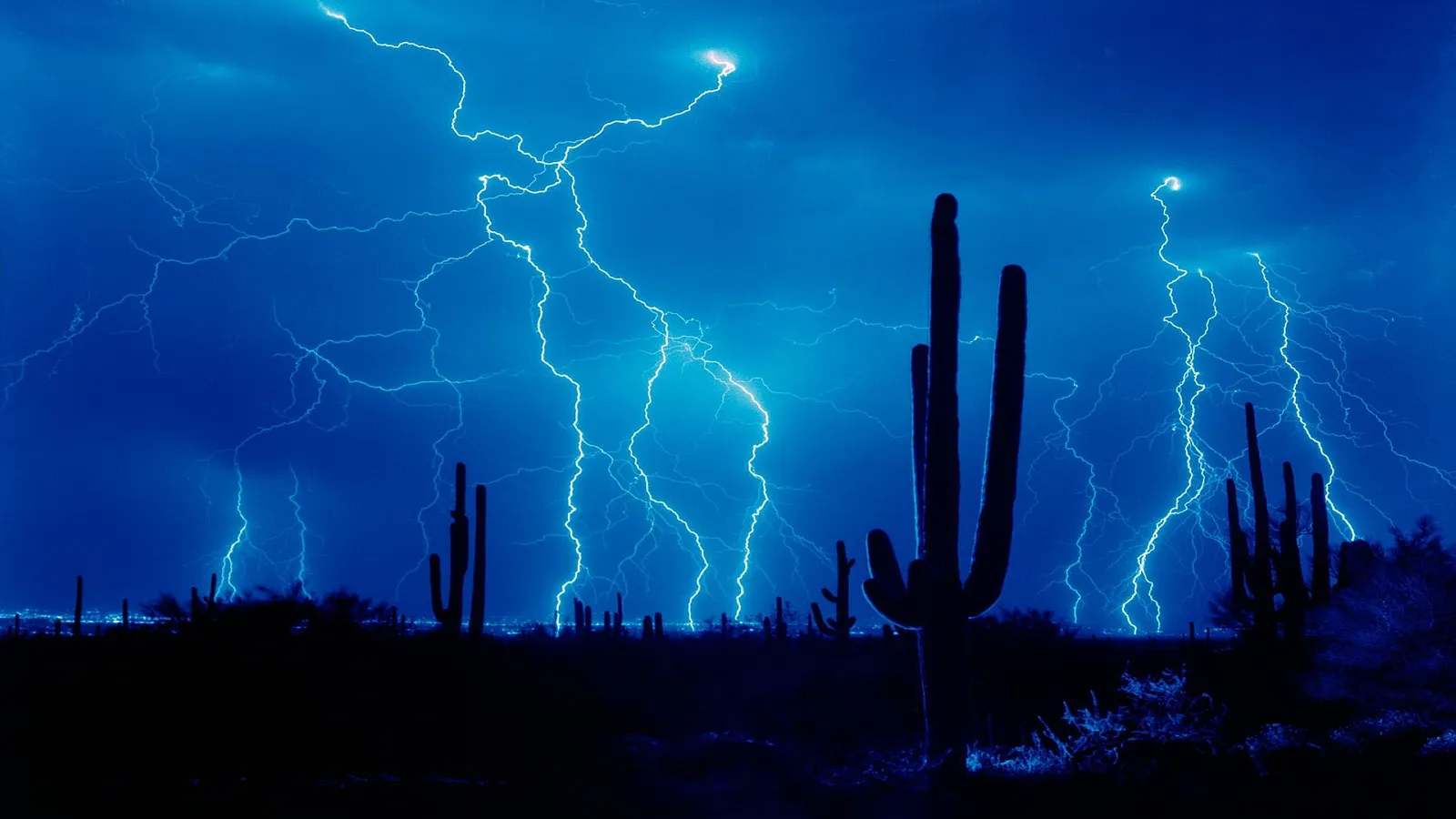 Jagged lightning bolts illuminate a dark desert landscape with silhouetted saguaro cacti.