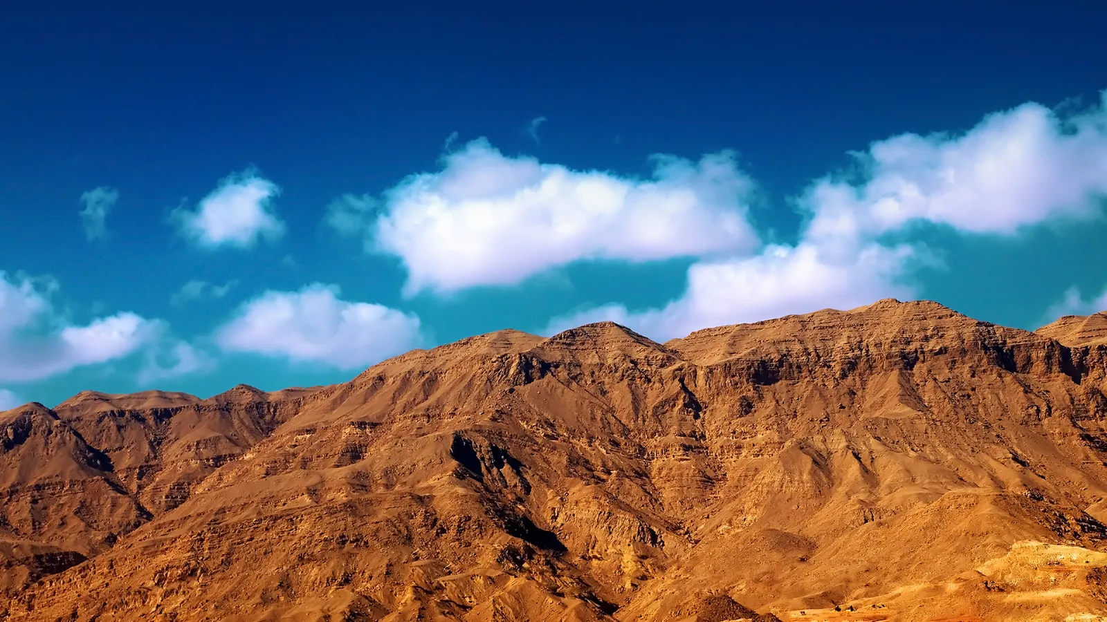 A wide view of rugged, brown desert mountains under a vibrant blue sky with scattered white clouds.