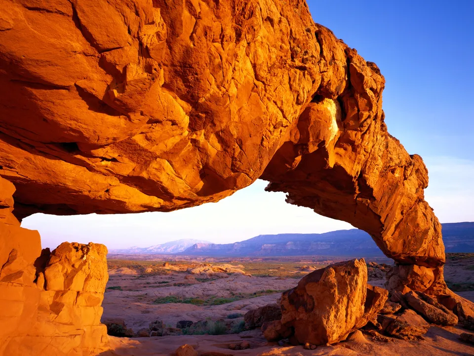 A large sandstone arch glows orange in the warm light of sunset, with a desert landscape stretching to the horizon.