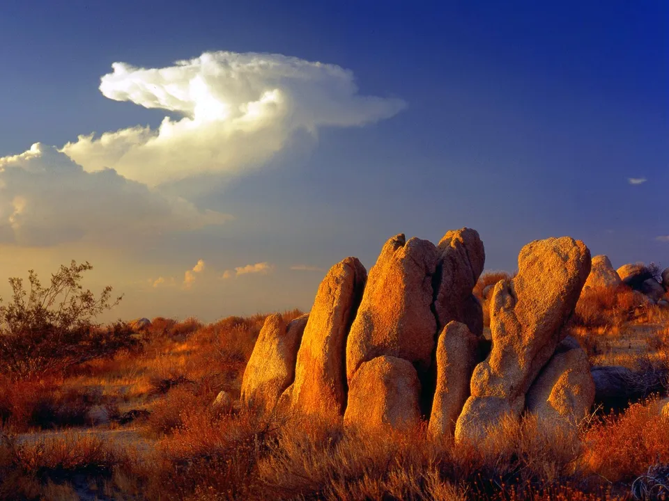 Large, weathered rock formations glow in the warm light of sunset, with dry brush and a dramatic cloud formation in the sky.