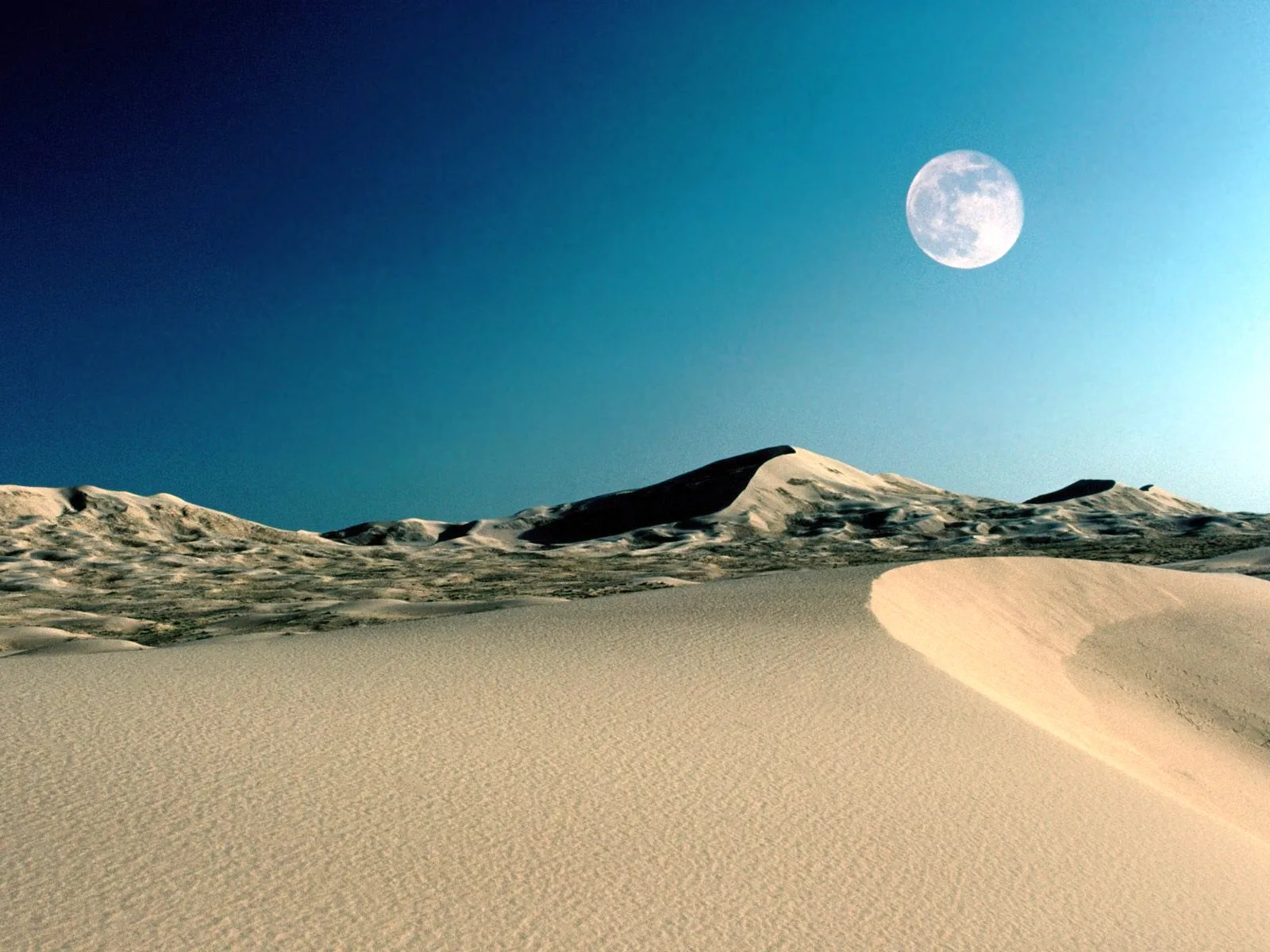 A full moon hangs in a clear blue sky above rolling sand dunes in a desert landscape.