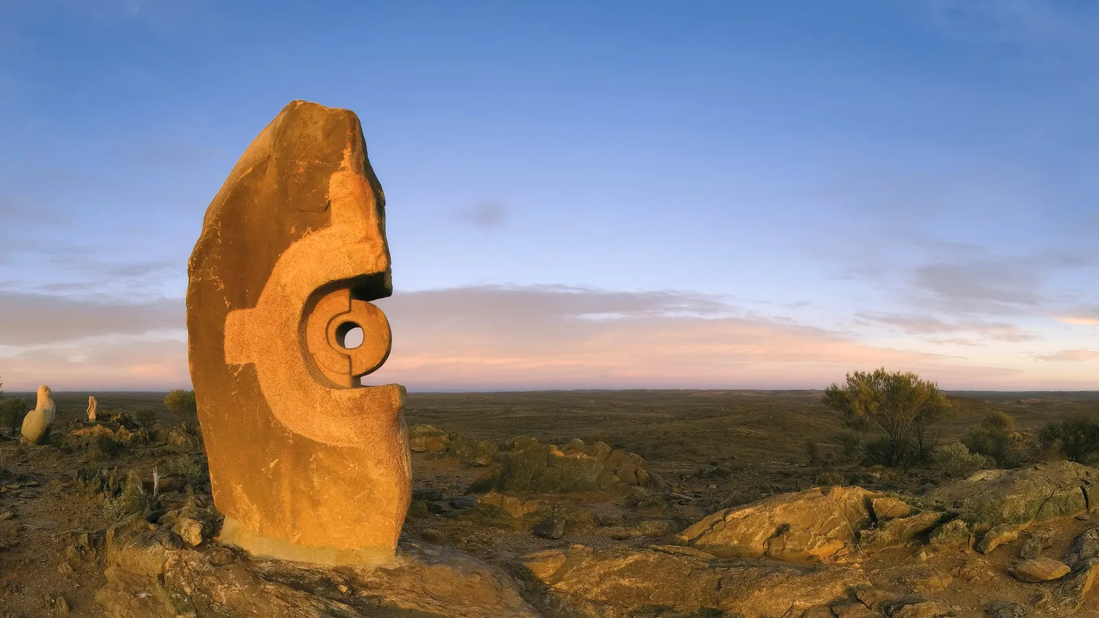 A large, carved stone sculpture stands in a barren desert landscape under a soft sunset sky.