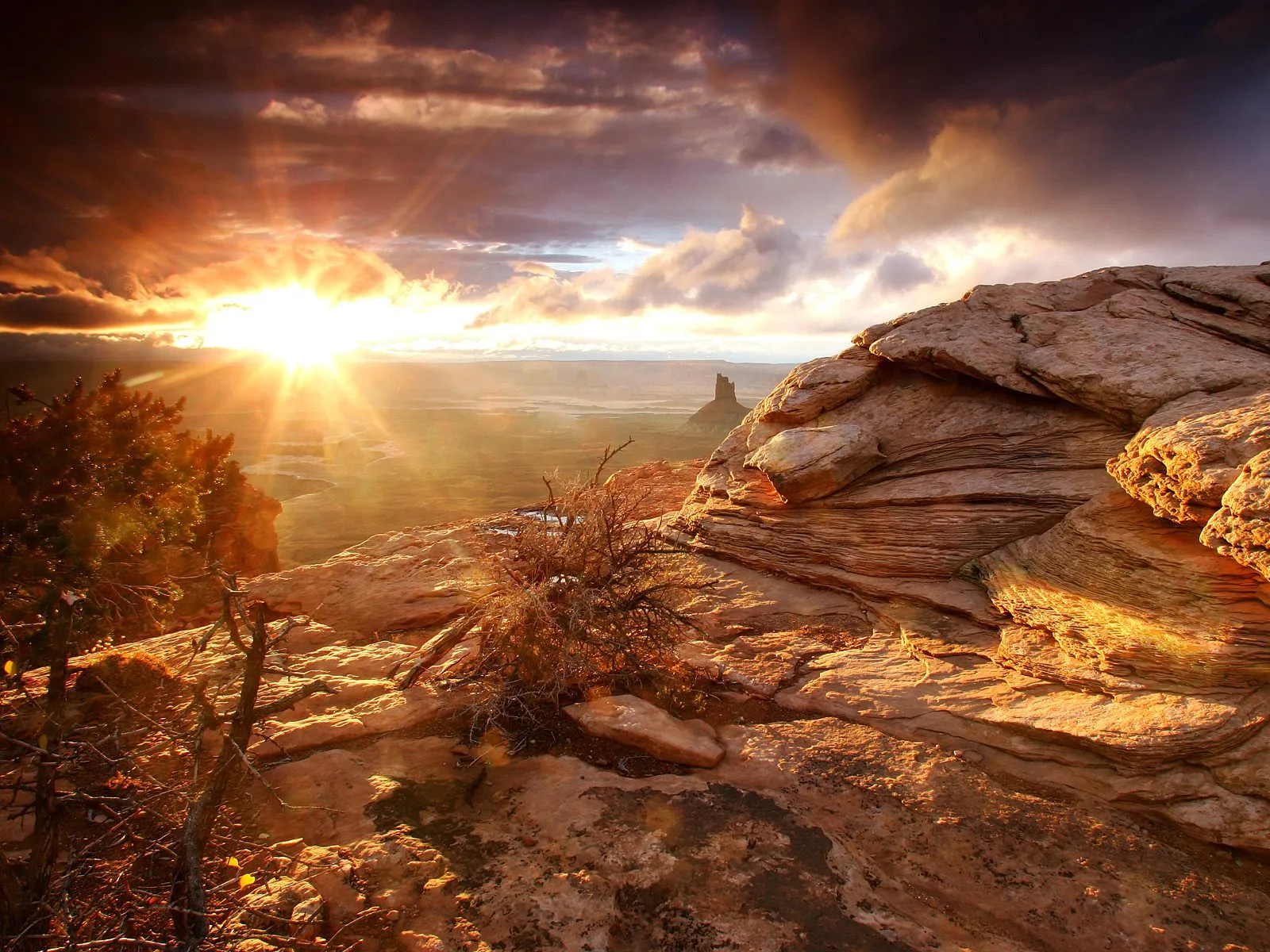 A brilliant sunburst rises over a vast desert canyon, illuminating rocky cliffs and sparse vegetation.