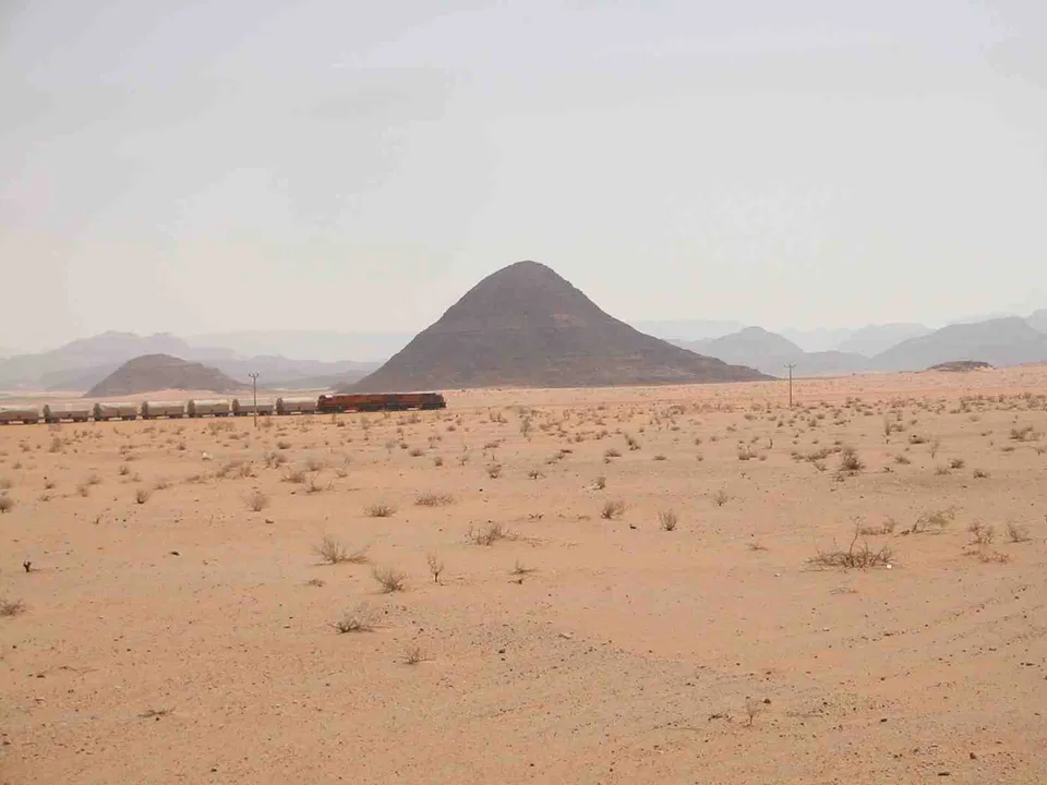 A long train travels across a sandy desert with mountains in the background under a hazy sky.