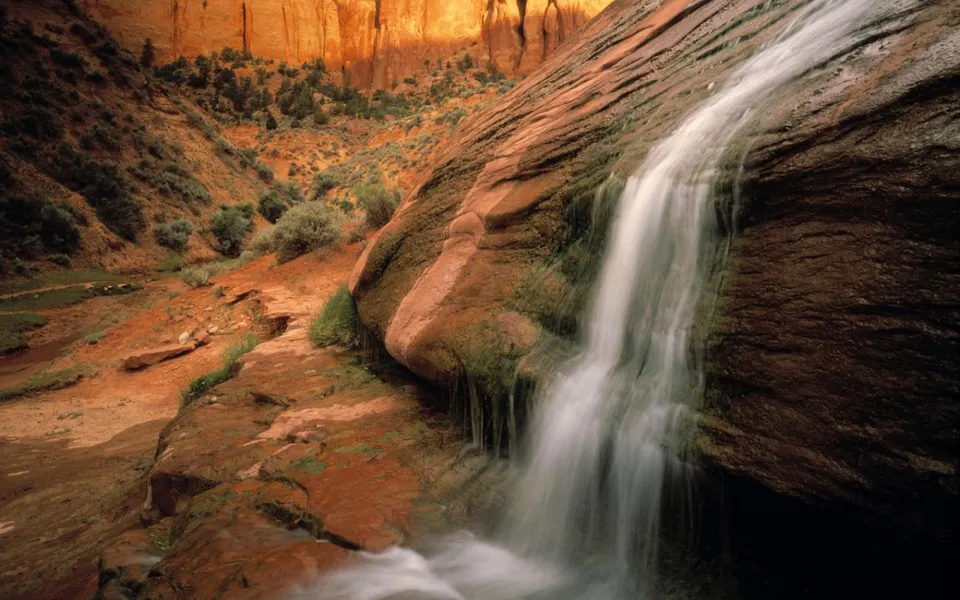 A thin waterfall flows down a large, textured red rock face in a desert canyon.