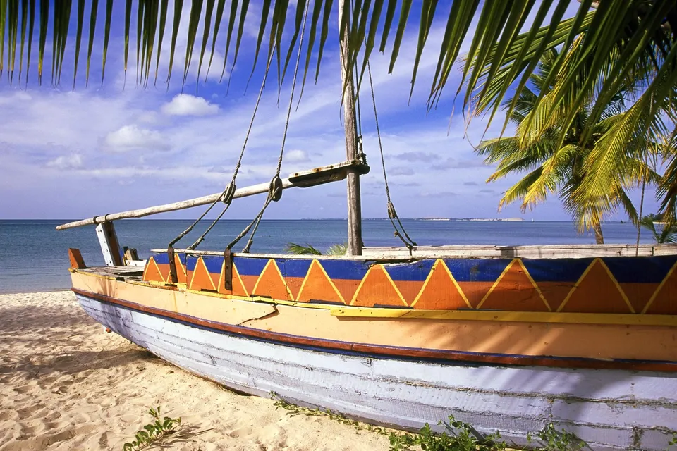 A colorful dhow boat rests on a sandy beach, with palm fronds framing the scene and the ocean in the background.